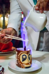 Purple flames rise during the creation of a Kona coffee in a bold tiki mug.