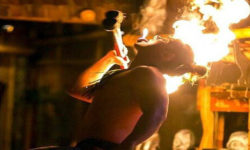 A close up of a Polynesian Dancer back lit by the high rising flames of fire torches held across his bare shoulder.