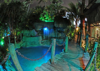 The romantic Mai-Kai boardwalk curving around the lagoon and waterfall illuminated in blue and green lights against the dark night sky.