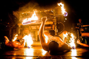 A close up of 2 Polynesian Dancers lit by the high rising flames of fire torches held under their knees.